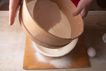 Large bowl, cutting board, sieve for sifting flour on the table. Female hands sifting flour in the home kitchen.