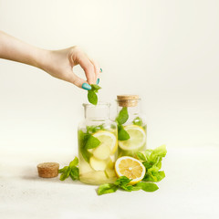 Female hand with green manicure making healthy drink lemonade in bottles with lemon, ginger and mint leaves at light background. Summer beverages making. Preparation of infused water with fruits