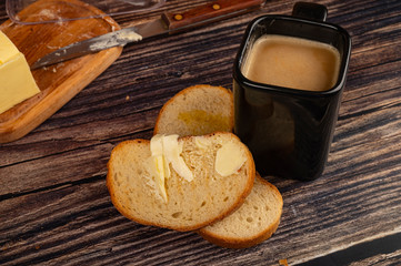Fresh wheat toast with butter, a ceramic mug with coffee and milk and a piece of butter in a wooden oil can on a wooden background. Close up.