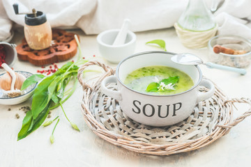 Green wild garlic soup in bowl with spoon on light table with ingredients. Close up. Seasonal home cuisine. Vegan food. Healthy cooking and eating