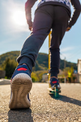 A teenage boy rides a scooter, pushing off with his left foot. Legs close-up. Bottom and rear view. Empty street in Sunny weather in the background. Concept of youth activity, sports and recreation