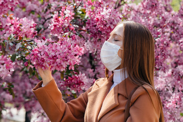 Fototapeta premium A young girl takes off her mask and breathes deeply after the end of the pandemic on a Sunny spring day, in front of blooming gardens. Protection and prevention covid 19