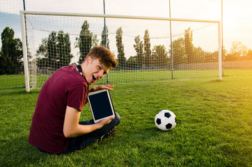 Naklejka premium Ecstatic boy holding tablet while sitting on the grass of football stadium 