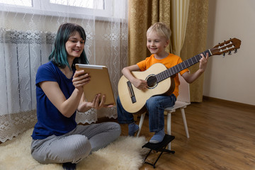 Cute boy plays the guitar, mom holds a tablet and shows to his son and smiles. A fun online lesson on the internet. Digital education in self isolation mode. Modern lifestyle. Stay at home concept.