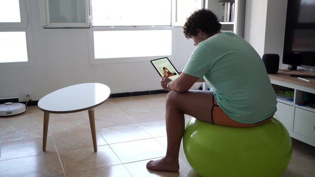 4k 24fps video of a young man with curly hair on a light blue shirt and orange shorts reading fake news on a online newspaper on his tablet in his living room while sitting on a green fit ball