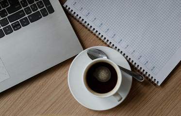 On the table are a cup of coffee with spoon, notebook, and a laptop.