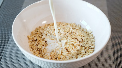 person pours fresh milk into ceramic bowl with tasty porridge cereals on grey table in hotel restaurant extreme close view