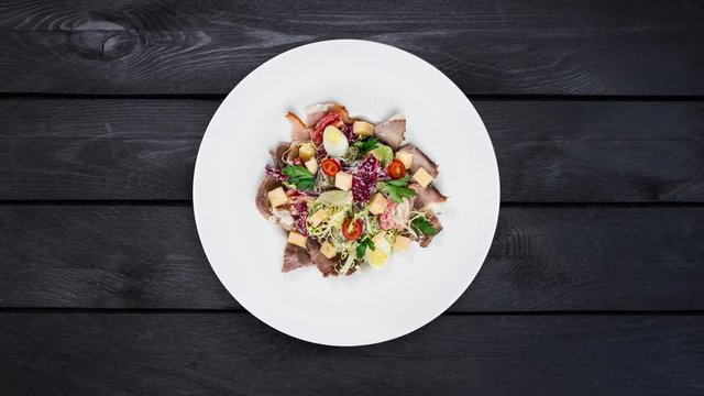 Rotating salad on a plate. Mixed lettuce, tomatoes, turkey smoked, smoked duck, dried horsemeat. Is at a black wooden background. Top view