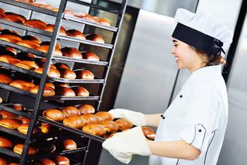 a young attractive woman Baker in a white chef's jacket and cap holds a baking tray with fresh hot rolls and bread on a shelf against the background of a bread factory or bakery. 