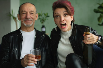 Indoor studio shot of excited beautiful woman with dark pink hair in white sweater and black leather jacket drinking with her senior father in loft style room. Female alcoholism