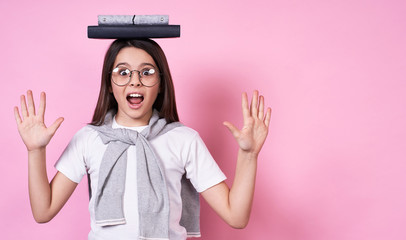 Beautiful young funny girl in glasses holds books on her head on a pink background.