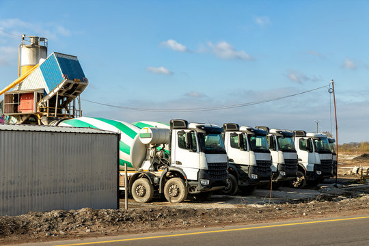 Row of many modern big mixer trucks parked against mobile temporary concrete plant factory at new asphalt road construction site morning day. Heavy machniery and industrial facilities background