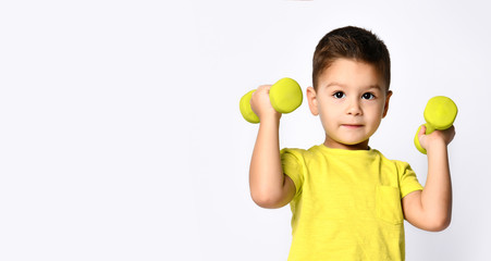 Little male in yellow t-shirt and denim shorts. He smiling, lifting dumbbell, posing isolated on white studio background