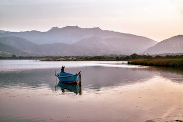 A boat on the lake before sunrise. Beautiful morning landscape, foggy mountains in the background. Sardinia, Italy.