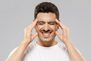 Young male experiencing severe headache, pressing fingers to temples, keeping eyes closed trying to ease pain, feeling sick and troubled, isolated on gray background