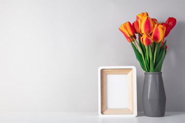 Mockup white photo frame on a white table and a wall. A vase of green flowers next to the frame. Copy space of a white wall.