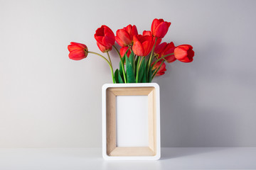 Mockup white photo frame on a white table and a wall. A vase of green flowers next to the frame. Copy space of a white wall.