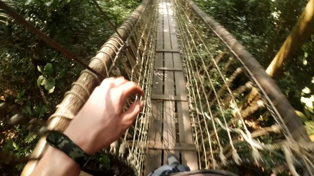 Man goes over the hanging bridge in Thailand jungle. First-person view of the feet stepping at the woods hanging above the trees. Theme of extremal activities at the vacation.