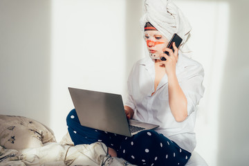 Young woman is working from home and her bed using laptop and phone during self isolation. She is wearing towel on her head and funny face mask. Early morning routine, online work, distance job