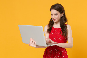 Smiling young brunette woman girl in red summer dress posing isolated on yellow wall background studio portrait. People emotions lifestyle concept. Mock up copy space. Working on laptop pc computer.