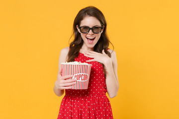 Laughing young woman girl in red summer dress, 3d glasses isolated on yellow background. People lifestyle concept. Mock up copy space. Watching movie film, hold bucket of popcorn, put hand on chest.