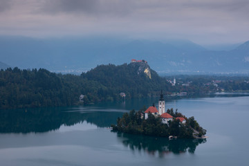 Church of pilgrimage Mariä Himmelfahrt on island in Lake Bled, during cloudy sunrise, castle in...