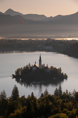 Church of pilgrimage Mariä Himmelfahrt on island and hot-air balloon at Lake Bled, during sunrise, sunbeams over mountains, Bled Slovenia.