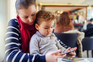 Portrait of small caucasian boy little child sitting in lap of his mother female woman by the table at home or cafe restaurant in day side view mobile smart phone playing games learning teaching app