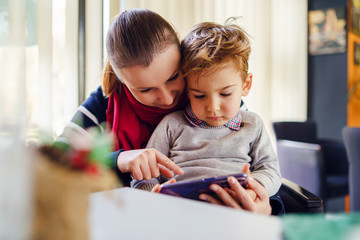 Portrait of small caucasian boy little child sitting in lap of his mother female woman by the table...