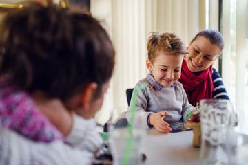 Obraz premium Small little boy caucasian child son sitting in the lap of his mother or aunt while female friend is looking at them teasing smiling holding smart mobile phone playing games making video call family