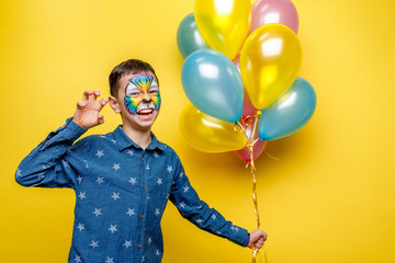 Emotional boy with aquagrim on birthday party, colorful tiger holding colorful balloons isolated on yellow background