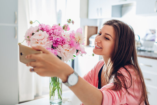 Woman taking selfie with bouquet of peonies flowers at home using smartphone. Gift present from husband