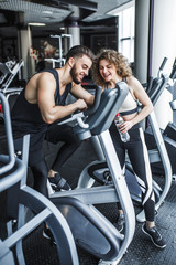 Fototapeta na wymiar A man and a woman are watching a training video. A group of two young people watching a video about exercise on a tablet. Man resting after workout sitting on the sports equipment in the gym