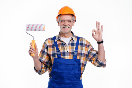 Photo of happy senior male builder being going to paint walls in house, holding paint roller in hand, wearing special uniform, apron and protective helmet, isolated on white background