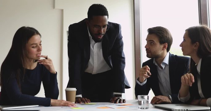 Young african american businessman in suit wearing eyeglasses, discussing project details with motivated teammates. Successful international company employees developing growth strategy in office.