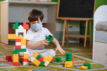 Little sad thoughtful bored toddler boy in mask playing colorful building blocks alone at home during quarantine. development game. Loneliness at self isolation period