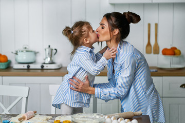 Little beautiful daughter kisses her mom in the kitchen while cooking sweets.