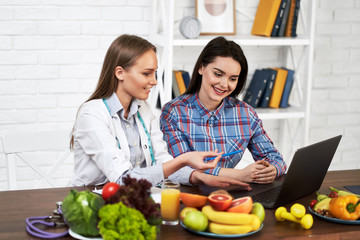 A smiling nutritionist advises a young patient woman on proper nutrition and dieting. The doctor shows in laptop a scheme of weight loss without a bad effect on health.