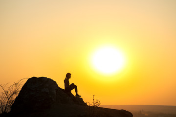 Silhouette of a woman hiker sitting alone on big stone at sunset in mountains. Female tourist on high rock in evening nature. Tourism, traveling and healthy lifestyle concept.