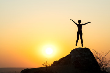 Fototapeta premium Silhouette of woman hiker jumping alone on empty rock at sunset in mountains. Female tourist raising her hands up standing on cliff in evening nature. Tourism, traveling and healthy lifestyle concept.