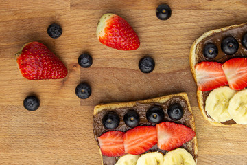 Top view on a toast bread with chocolate topping, banana, strawberry and blueberry on a wooden board. Fruit snack concept.