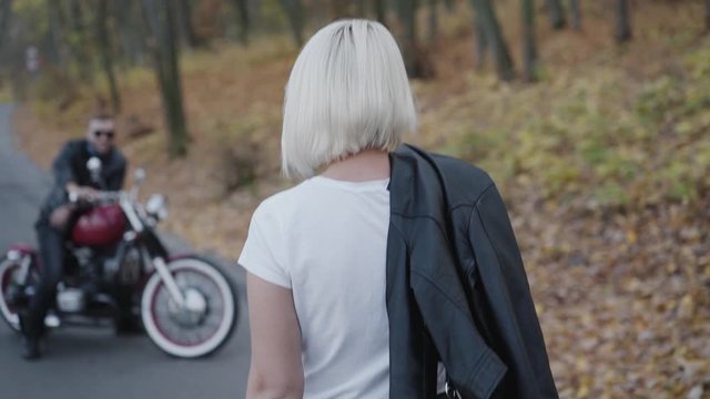 Back look of blonde comes to her smiling brutal biker with motorcycle in park