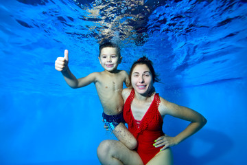 Obraz na płótnie Canvas A happy mother and her young son pose for the camera underwater in a pool on a blue background. Mom in a red knitted swimsuit looks at the camera. They smile. Closeup. Digital photo.