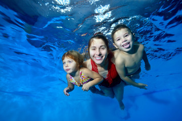Obraz na płótnie Canvas Sports family: a smiling mother and her two young children swim underwater in the pool. They hug, smile and pose for the camera on a blue background. Close up. Digital photo.