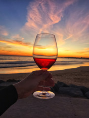 Woman man hand with a glass of red wine on a background of sunset sky and waves on the beach by the...