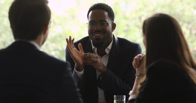 Happy young african american well-dressed businessman in eyeglasses talking to diverse partners in office. Smiling biracial manager negotiating business ideas project details with colleagues.