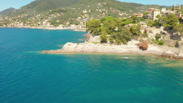 Fly over the rocky sea coast with a white statue of Jesus on the top and mountains on the background. Traditional italian colorful housees on the hill. Huge stones under the water Camogli, Italy.