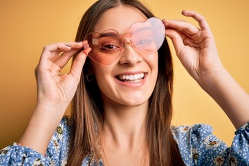 Young beautiful brunette woman wearing funny sunglasses with heart over yellow background with a happy face standing and smiling with a confident smile showing teeth