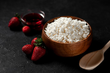 tasty fresh homemade cottage cheese in a wooden bowl with strawberries and jam is on a dark background, healthy food on a black table