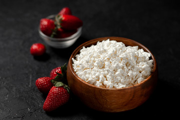 homemade fresh cottage cheese in a wooden plate without additives lies with strawberries on a black texture table